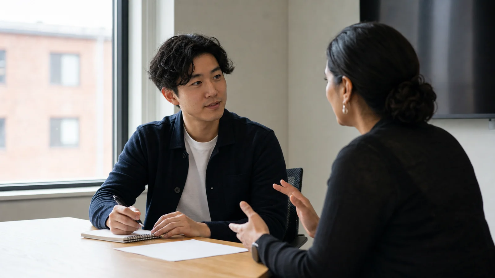 Realistic editorial photograph of a new graduate discussing constructive feedback with a workplace supervisor