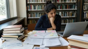 Realistic editorial photograph of an international student narrowing a research topic with notes and library sources