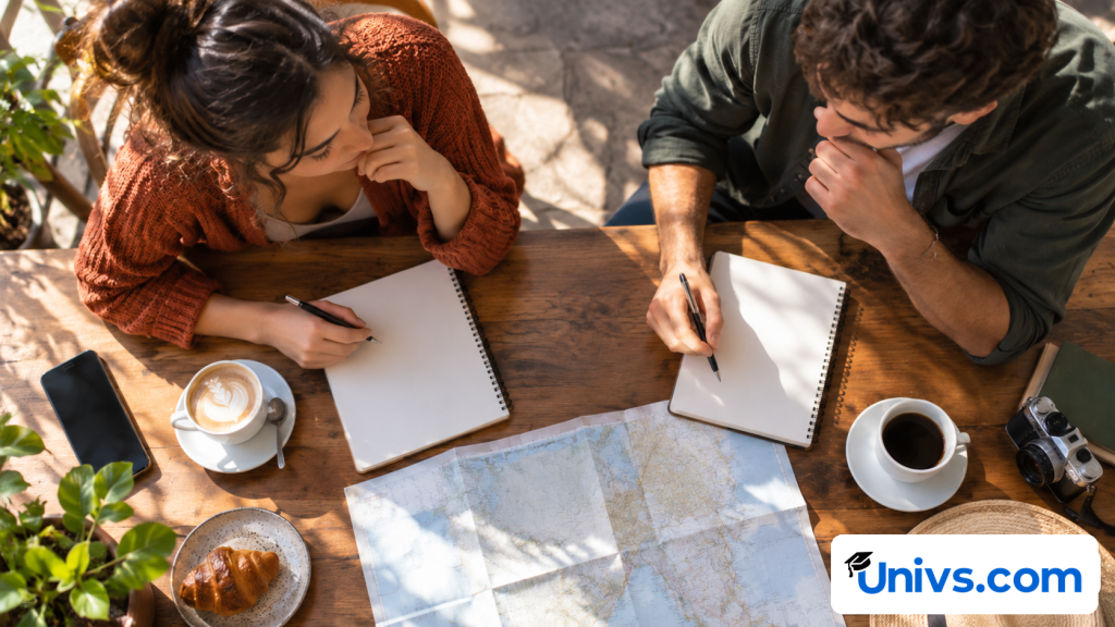 Two university-age students comparing two study destinations using two blank notebooks at a sunny cafe table; overhead view with hands, warm coffee cups and a folded blank map